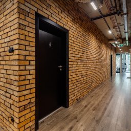PRIMATER SPÓŁKA Z OGRANICZONĄ ODPOWIEDZIALNOŚCIĄ - Korytarz biurowy z ceglanymi ścianami, drewnianą podłogą i czarnymi drzwiami z symbolem toalety damskiej.