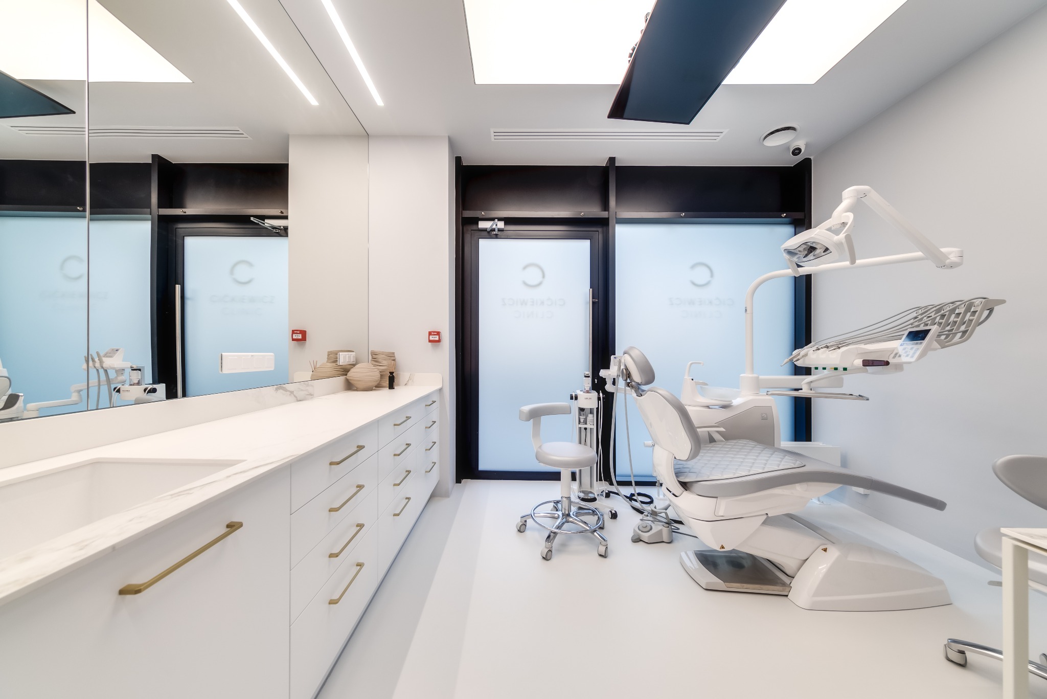 Nowoczesny gabinet stomatologiczny z białymi meblami, marmurowym blatem i złotymi uchwytami, oświetlony podłużnymi lampami LED.