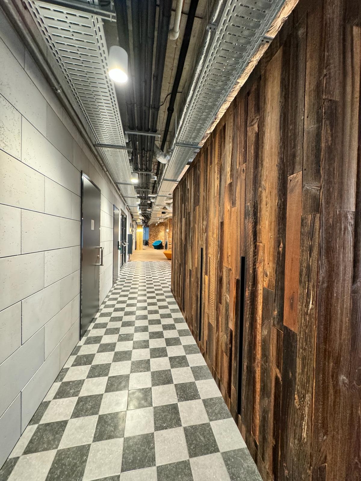 Długi korytarz w biurze z szachownicową podłogą, wykończony drewnianą ścianą z desek o różnej fakturze i jasną, kafelkową ścianą po drugiej stronie, z widocznymi instalacjami pod sufitem.