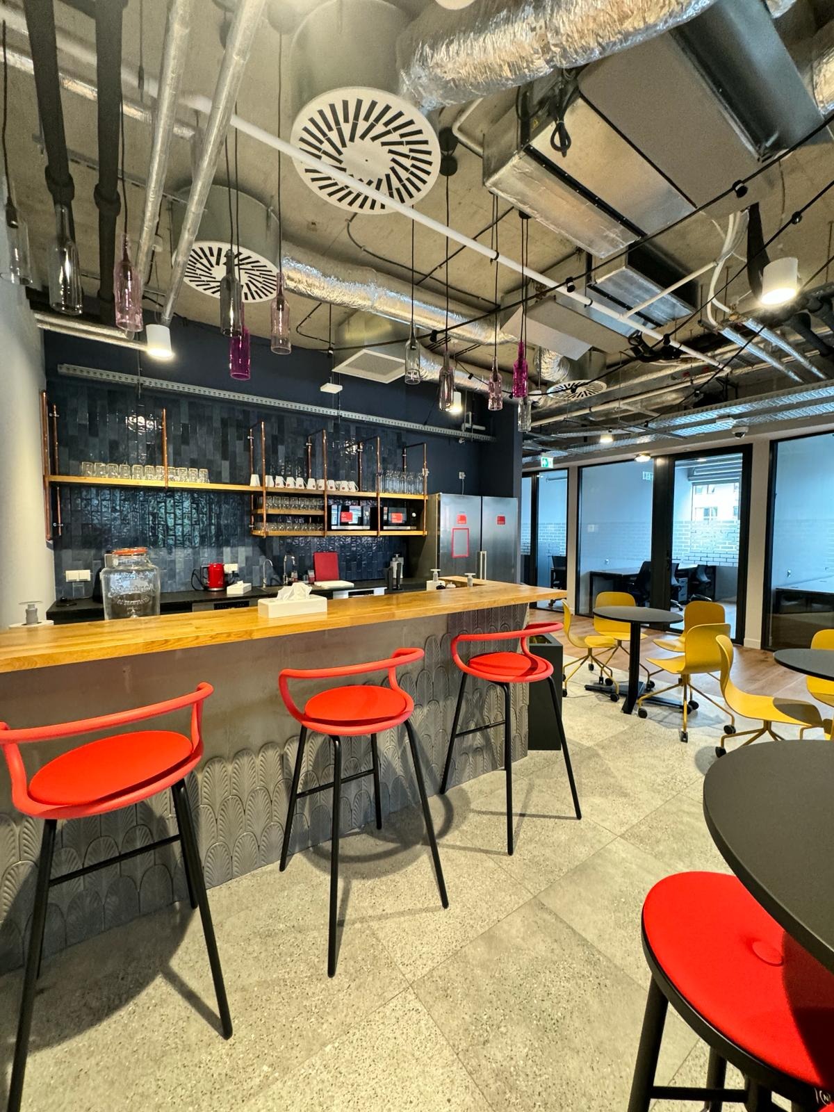 Nowoczesna przestrzeń biurowa z barem: drewniany blat, czerwone hokery, odsłonięte instalacje sufitowe i przeszklone ściany działowe.