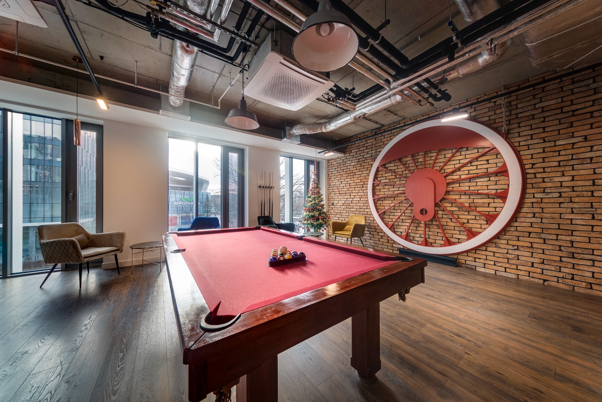 Pokój biurowy z betonowym sufitem i widocznymi instalacjami, ceglaną ścianą z dekoracją w kształcie koła, stołem bilardowym i choinką w rogu.
