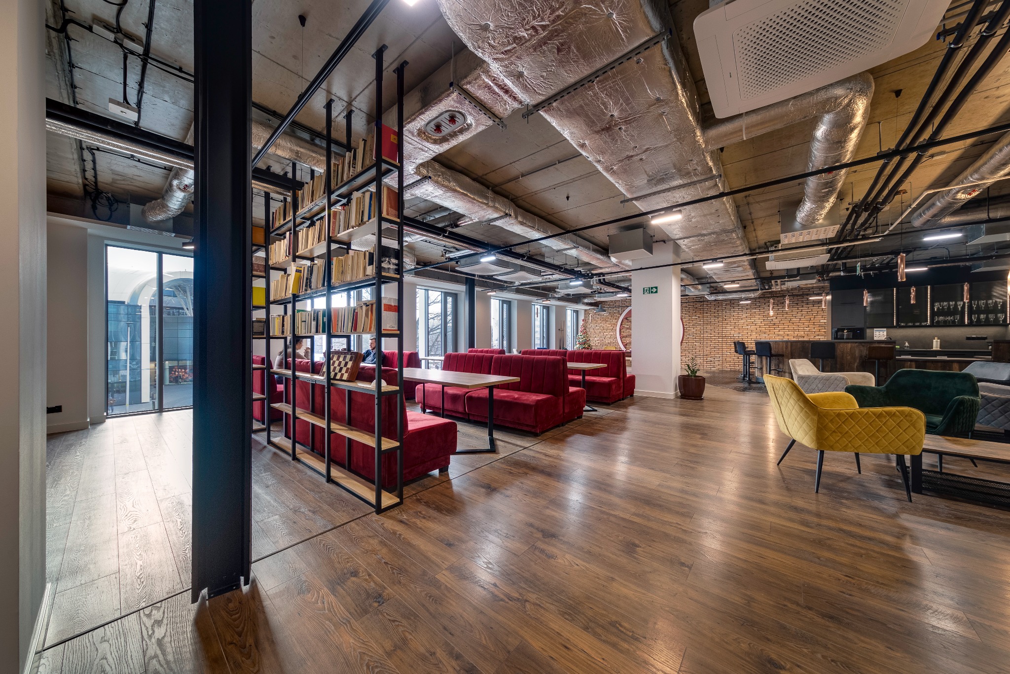 Przestronne wnętrze biurowe z drewnianą podłogą, widocznymi instalacjami sufitowymi, regałem z książkami i czerwonymi sofami w stylu retro, oraz strefą relaksu z barem i krzesłami barowymi w tle.