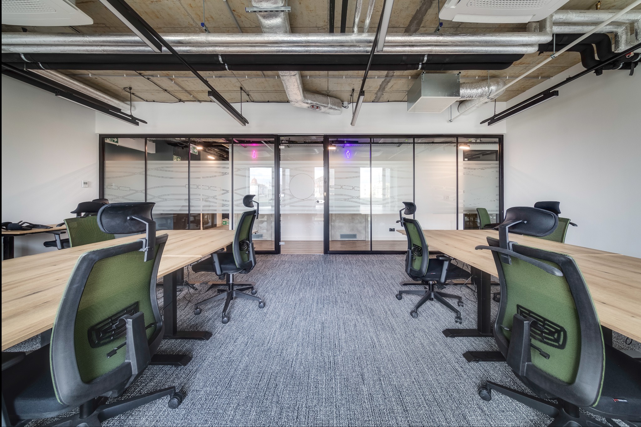 Nowoczesne biuro z długimi drewnianymi biurkami, ergonomicznymi krzesłami z zielonymi oparciami i szarą wykładziną dywanową. Szklane ścianki działowe z matowymi wzorami oddzielają przestrzeń,...