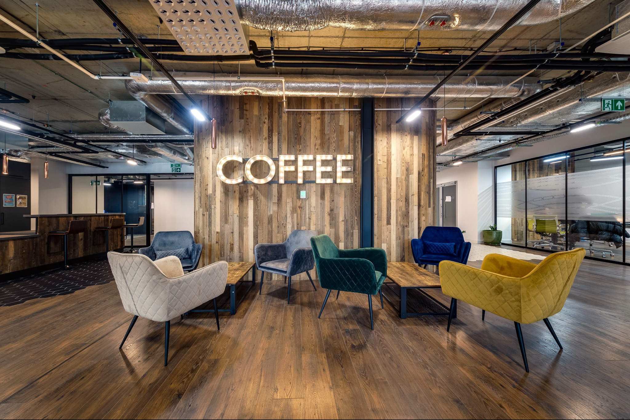 Nowoczesna przestrzeń biurowa z drewnianą ścianą z podświetlonym napisem 'COFFEE', różnokolorowe welurowe fotele i drewniana podłoga.