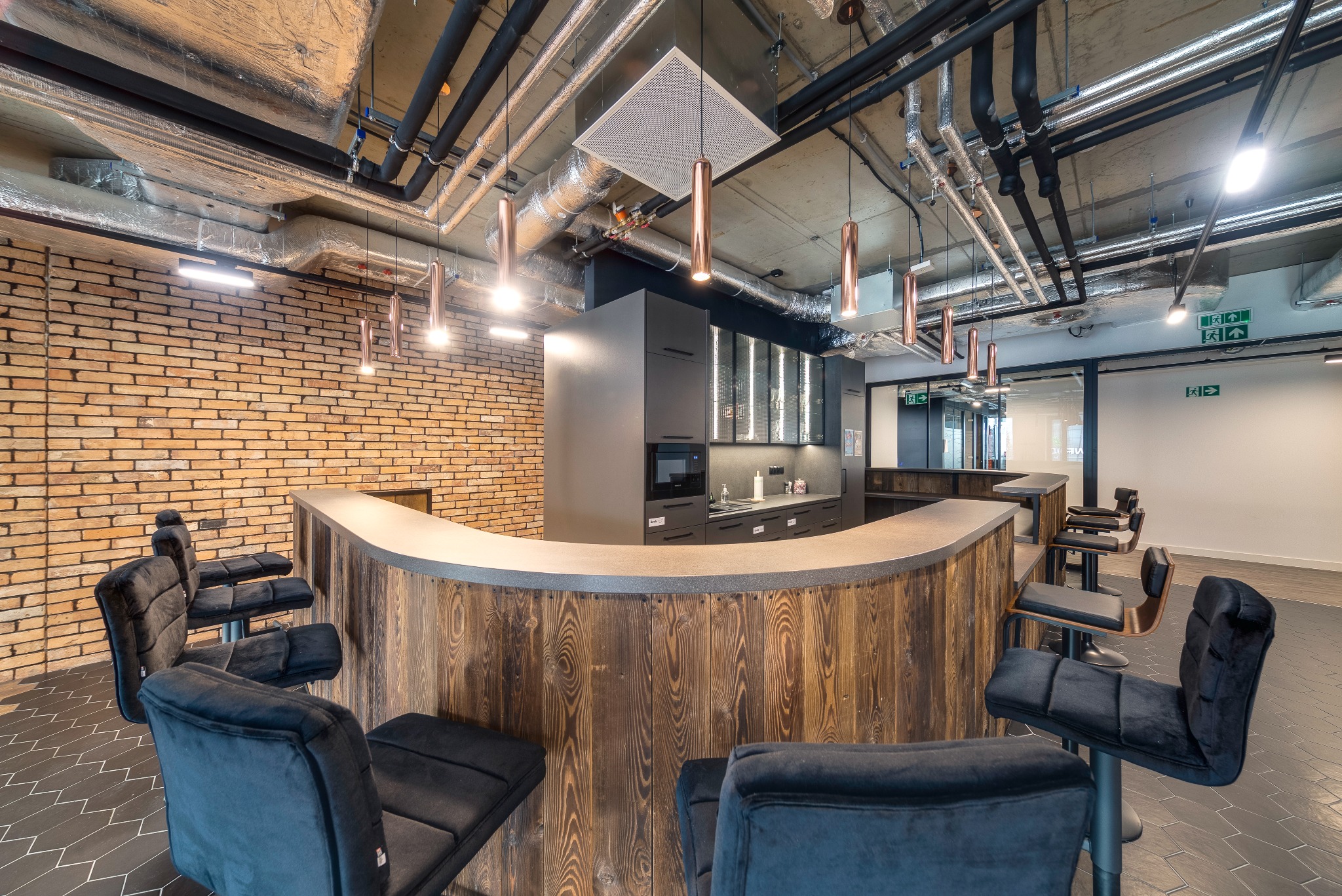 Nowoczesna przestrzeń biurowa z drewnianą ladą barową, welurowymi krzesłami, ceglaną ścianą i odsłoniętymi instalacjami na suficie, oświetlona wiszącymi lampami w miedzianym kolorze.