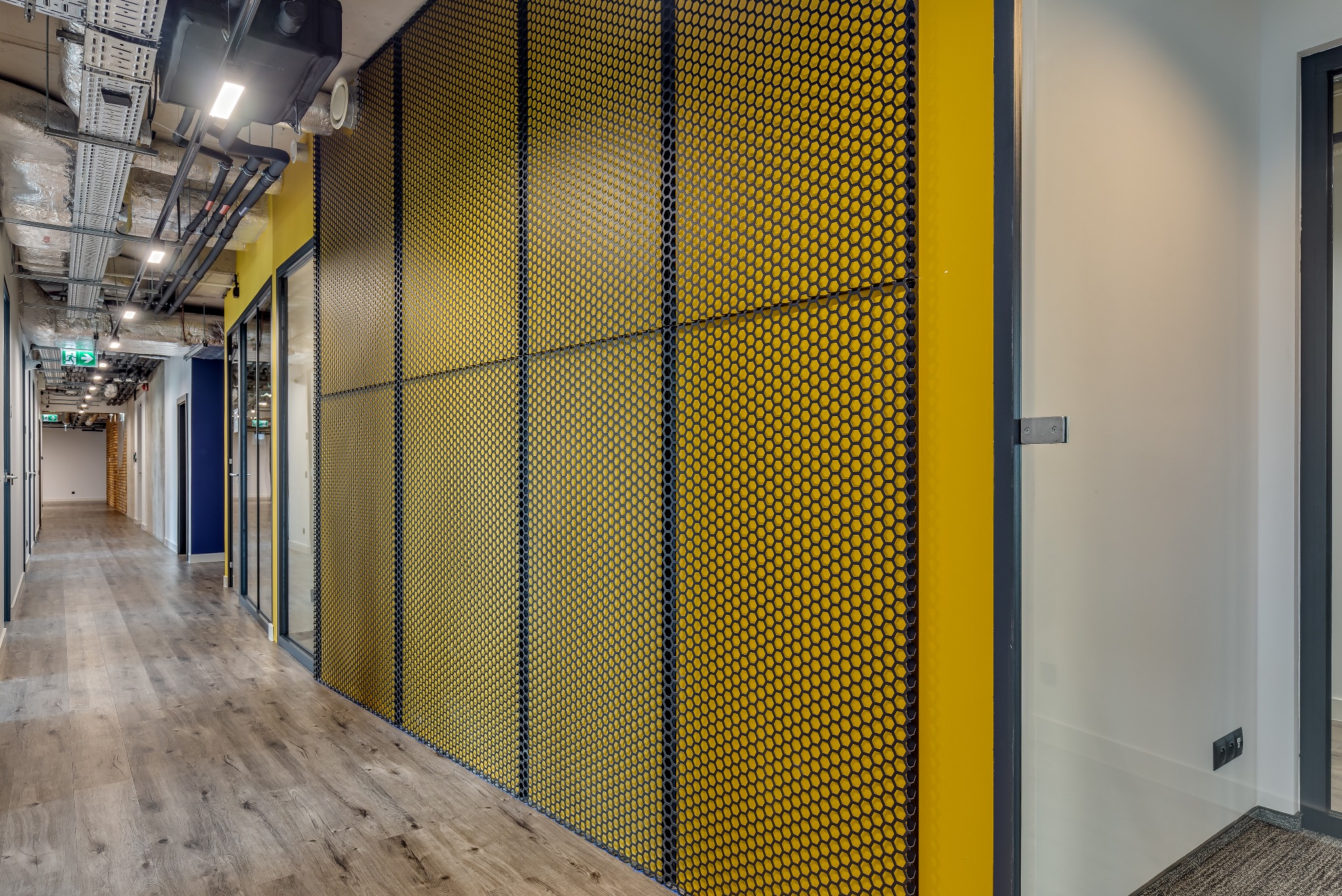 Korytarz biurowy z drewnianą podłogą, żółtą ścianą z metalową siatką o strukturze plastra miodu i odsłoniętą instalacją sufitową.