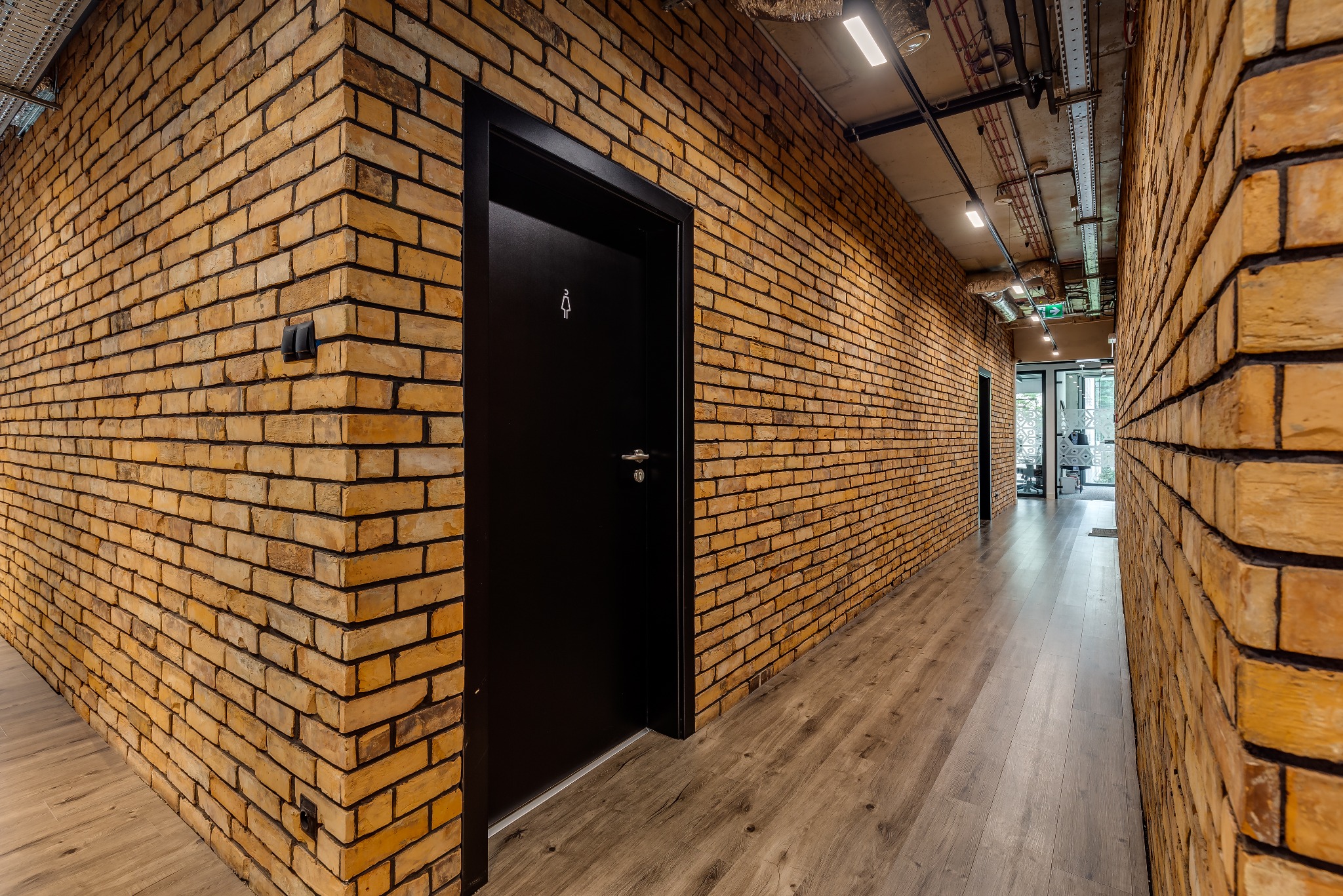 Korytarz biurowy z ceglanymi ścianami, drewnianą podłogą i czarnymi drzwiami z symbolem toalety damskiej.