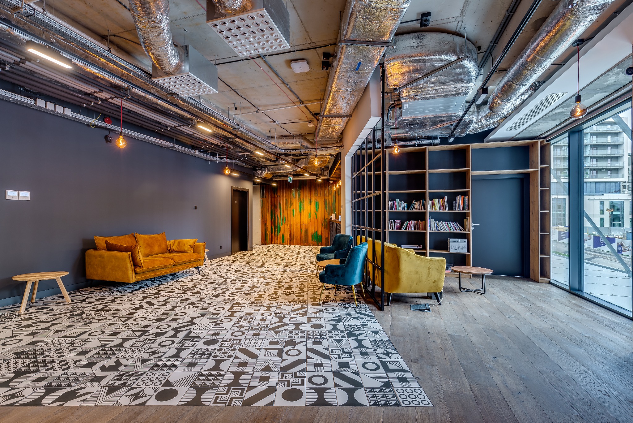 Przestronne biuro z wzorzystą podłogą, żółtą sofą, turkusowymi fotelami i widoczną instalacją wentylacyjną pod sufitem.