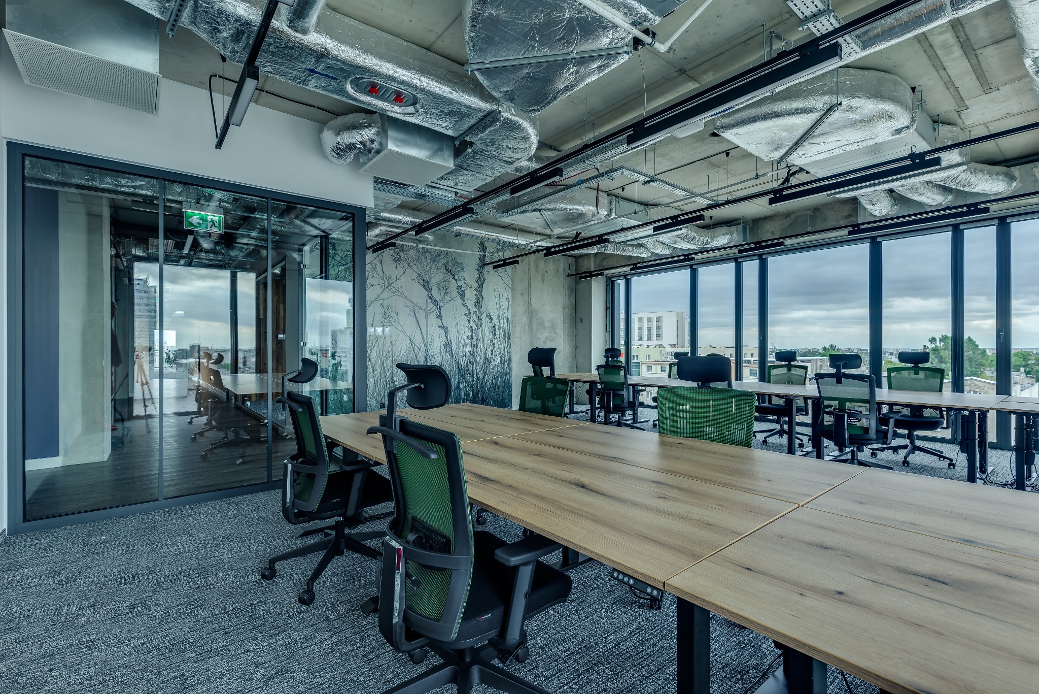 Nowoczesne biuro z długimi drewnianymi stołami i ergonomicznymi krzesłami z zielonymi akcentami, widok na miasto przez duże okna, surowy betonowy sufit z widoczną instalacją wentylacyjną.
