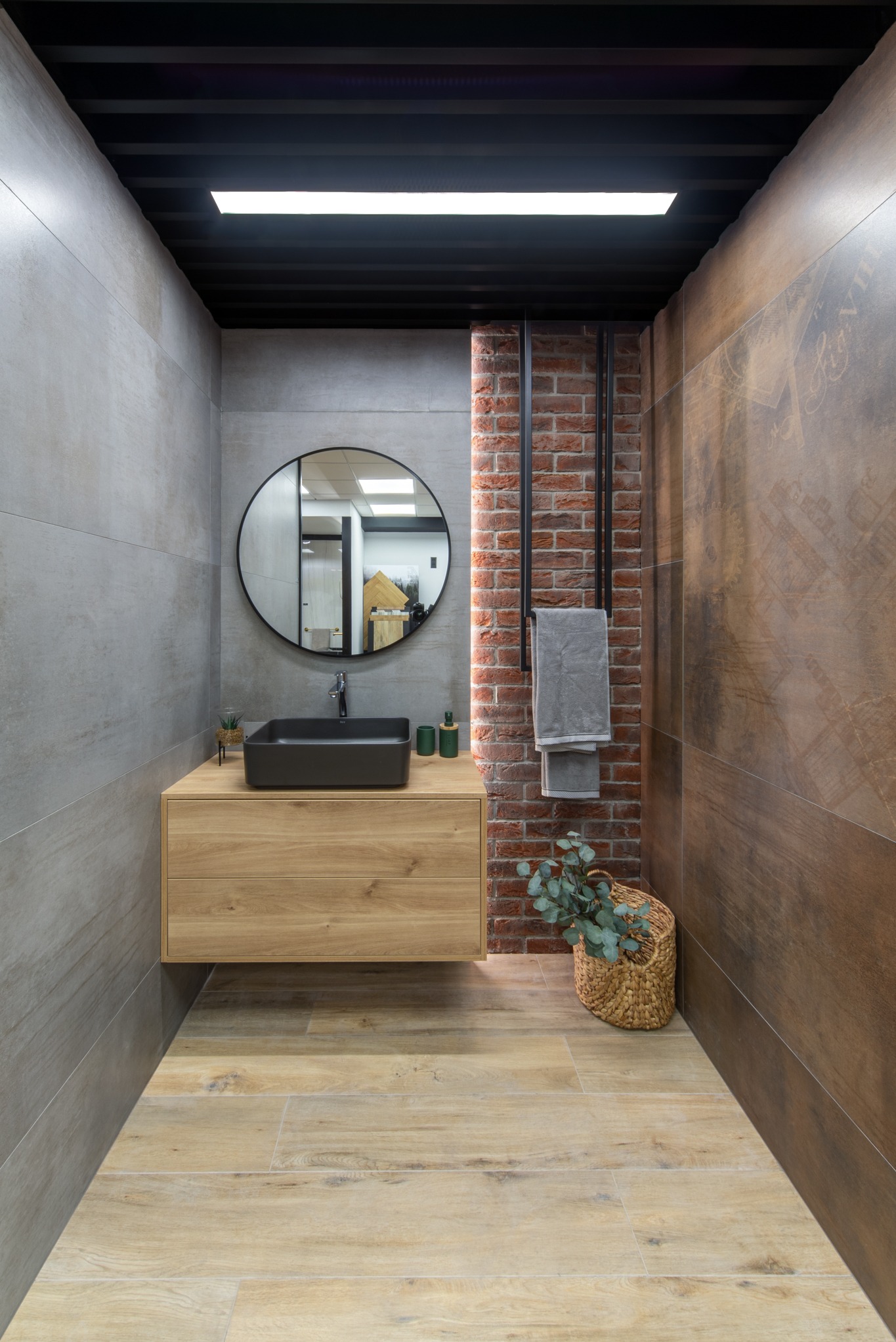 Nowoczesna łazienka z drewnianą szafką pod umywalkę, czarną umywalką i okrągłym lustrem na tle ściany z cegły i dekoracyjnych płytek. Wnętrze uzupełnia kosz z rośliną.