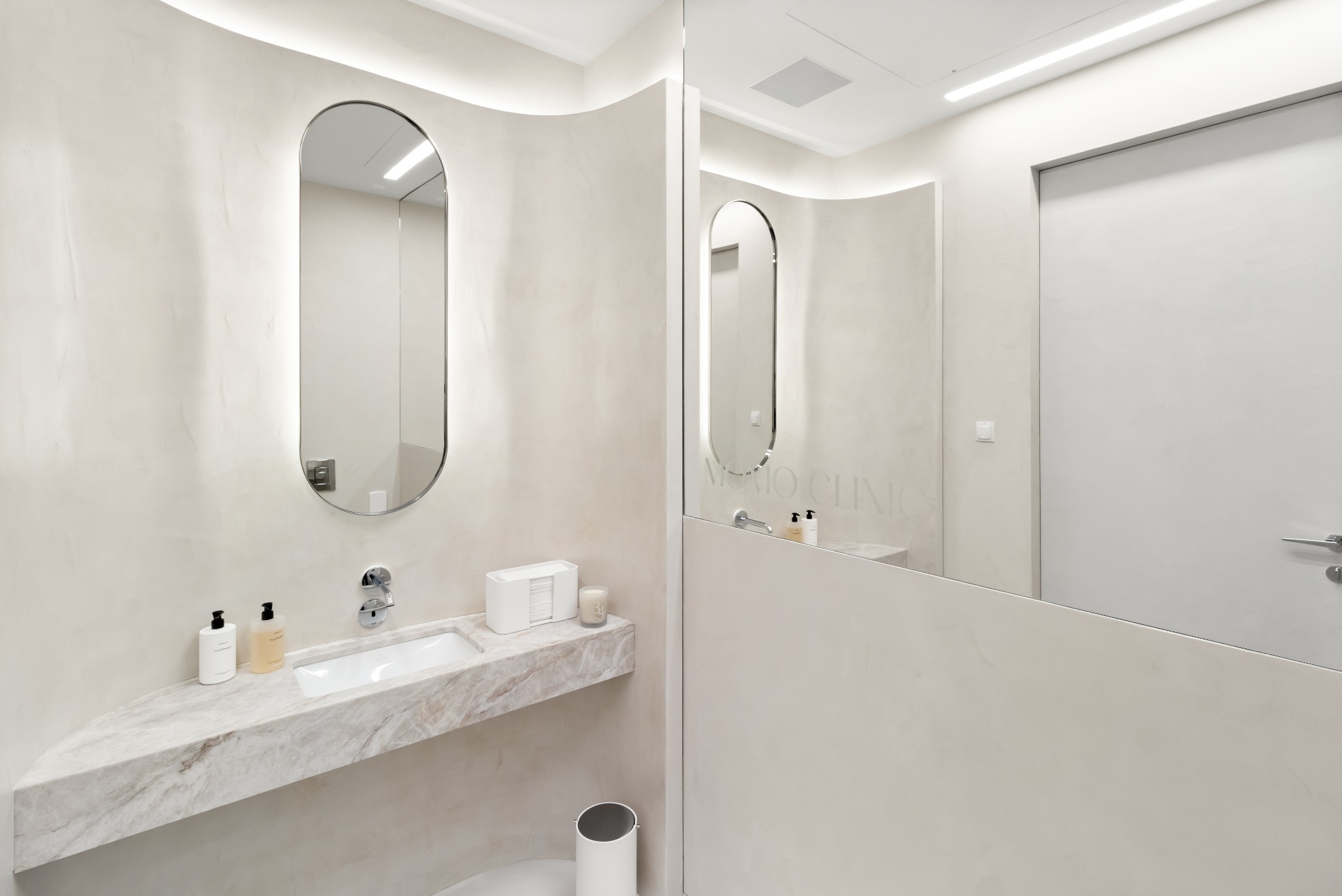 Nowoczesna łazienka w biurze z umywalką na marmurowym blacie, owalnym lustrem z podświetleniem LED, minimalistycznym dozownikiem mydła i dekoracyjną świecą zapachową.