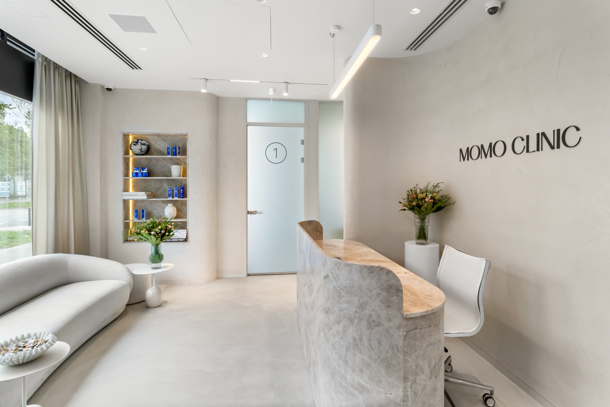 Jasne, minimalistyczne wnętrze biura kliniki MOMO z zakrzywioną ladą recepcyjną z marmuru, sofą w kształcie półksiężyca, dekoracyjną półką z oświetleniem i przeszklonymi drzwiami z numerem 1.