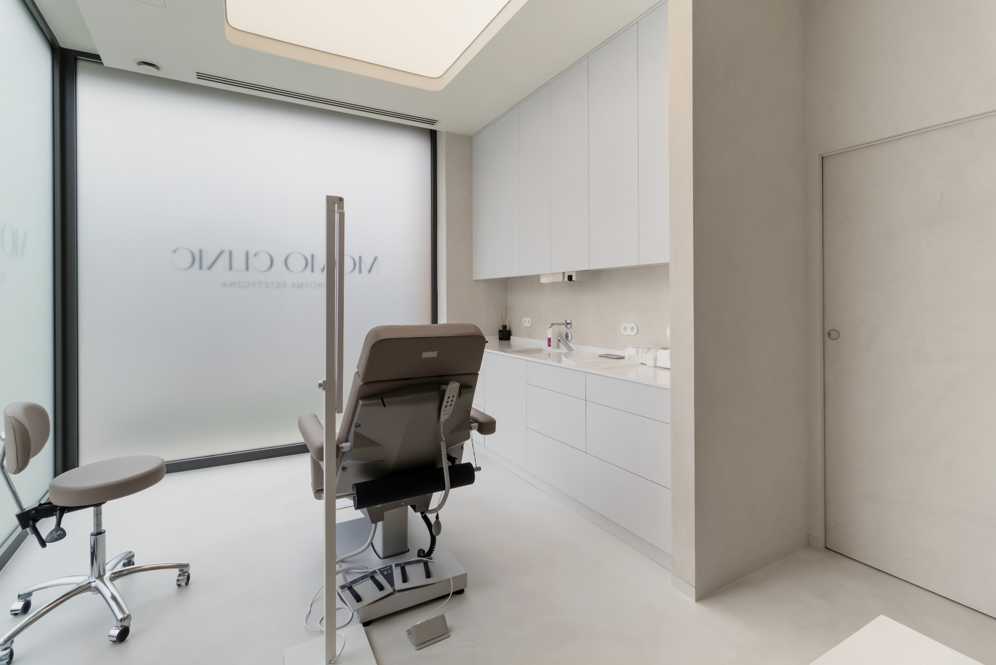 Jasne, minimalistyczne wnętrze gabinetu lekarskiego z fotelem zabiegowym, umywalką i zabudową meblową. Na matowej szybie okna widoczne logo 'VICINO CLINIC'.