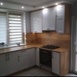 Tamro - Jasna kuchnia z białymi szafkami, drewnianym blatem i okładziną ścienną imitującą drewno, widok na ogród przez przeszklone drzwi balkonowe.