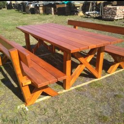 Tamro - Komplet drewnianego stołu z ławkami w kolorze czerwonego brązu, z charakterystycznym krzyżowym wzmocnieniem nóg, ustawiony na trawie przed stosem drewna i cegieł.