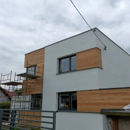 Tamro - Nowoczesny dom z elewacją częściowo wykończoną panelami drewnianymi, widoczne rusztowanie przy ścianie, szara metalowa barierka w tle.