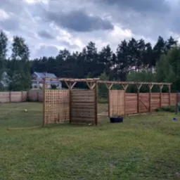 Drewniana pergola z kratownicowymi panelami i ogrodzeniem w tle, widok na posesję z domem pod pochmurnym niebem.