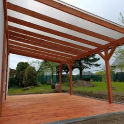 Nowoczesna konstrukcja z drewna i przezroczystego zadaszenia przylegająca do budynku, otoczona zielonym trawnikiem i drzewami.