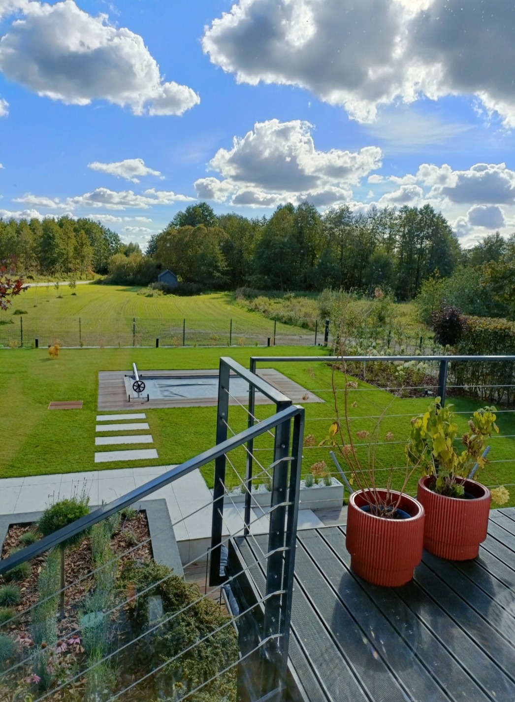 Nowoczesny taras z ciemnych desek kompozytowych, balustrada z metalowymi słupkami i linkami, widok na zadbany trawnik z basenem i drewnianym podestem w tle, błękitne niebo z obłokami.
