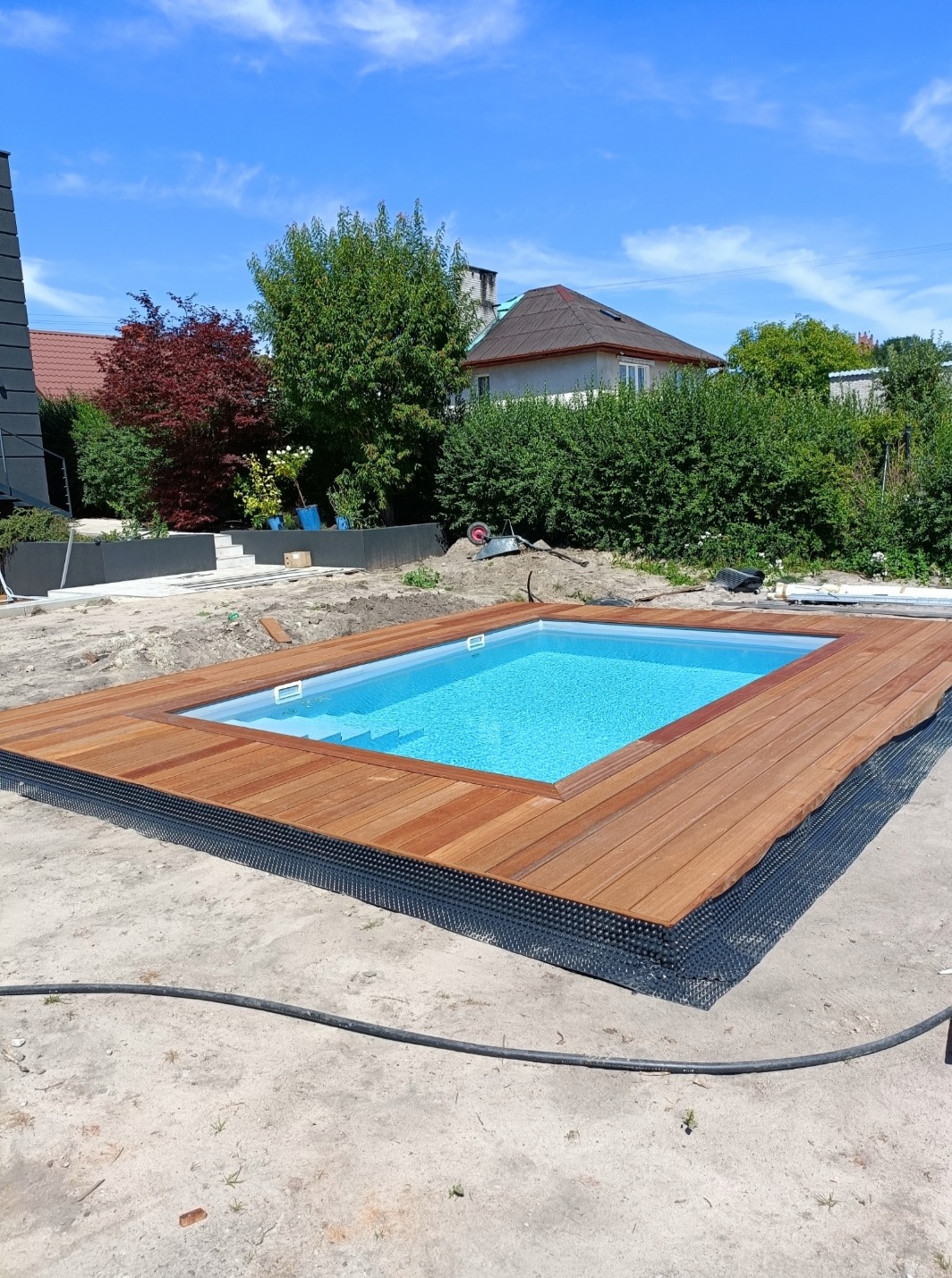 Basen z turkusową wodą otoczony nowym, drewnianym podestem na czarnej membranie, w tle domy i zieleń ogrodu.