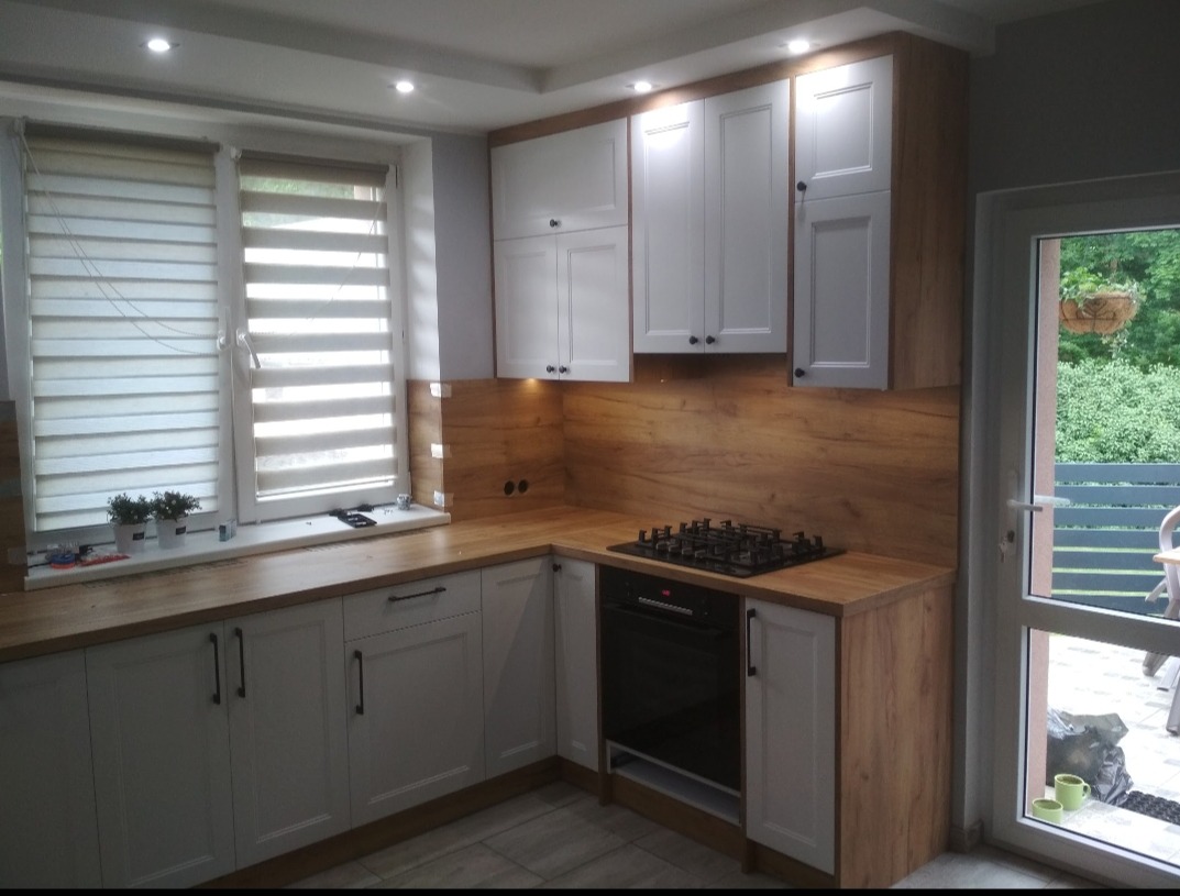 Jasna kuchnia z białymi szafkami, drewnianym blatem i okładziną ścienną imitującą drewno, widok na ogród przez przeszklone drzwi balkonowe.