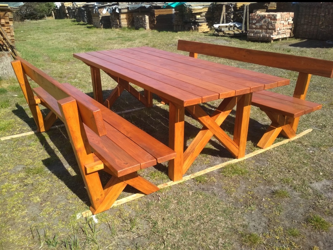 Komplet drewnianego stołu z ławkami w kolorze czerwonego brązu, z charakterystycznym krzyżowym wzmocnieniem nóg, ustawiony na trawie przed stosem drewna i cegieł.