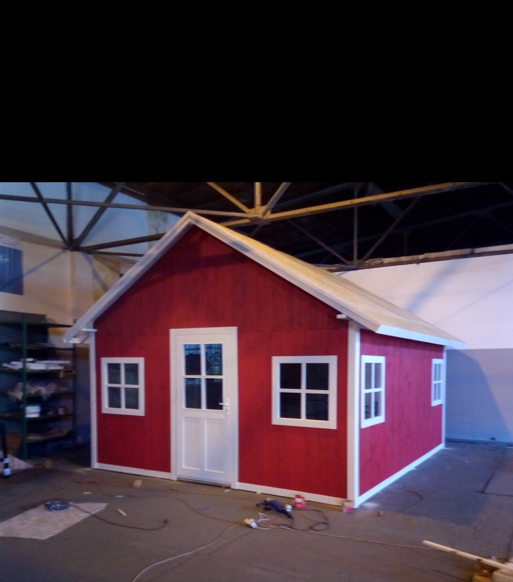 Czerwony domek dla dzieci z białymi ramami okiennymi i drzwiami, widoczny w hali produkcyjnej z narzędziami na podłodze.