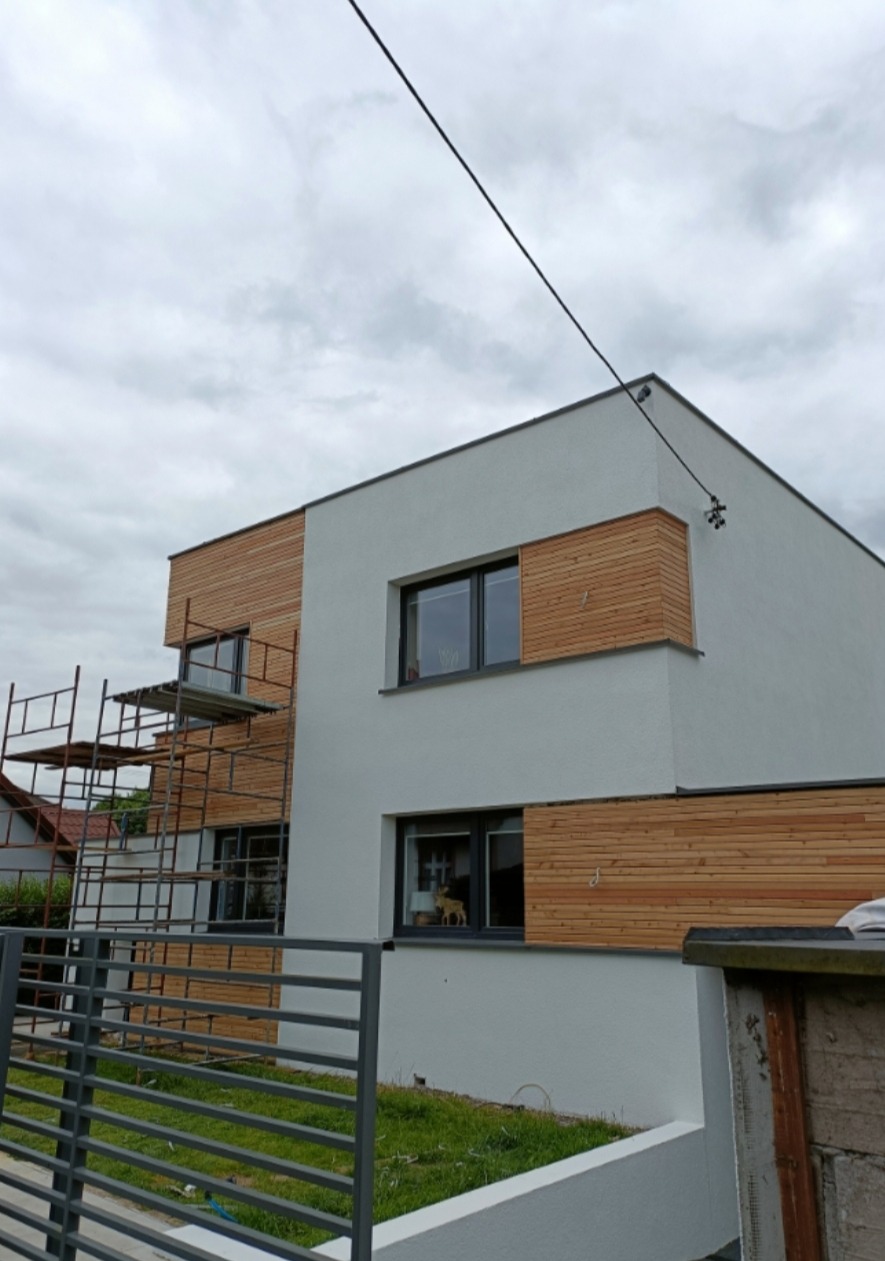 Nowoczesny dom z elewacją częściowo wykończoną panelami drewnianymi, widoczne rusztowanie przy ścianie, szara metalowa barierka w tle.