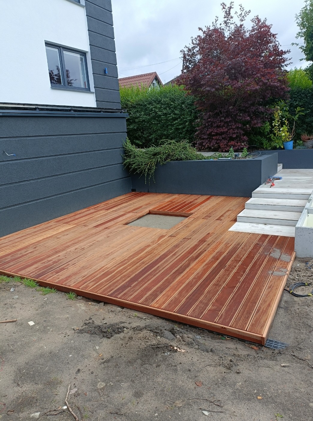 Częściowo ułożony taras z desek w kolorze ciepłego brązu, z kwadratowym otworem pośrodku, obok betonowych schodów i rabatki z zielenią, na tle nowoczesnego domu z szarą elewacją.