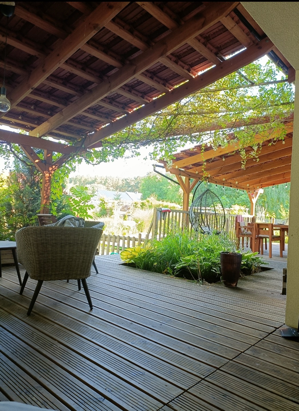 Zadaszony drewniany taras z roślinami w Karpinach, widok na ogród i domy w tle, rattanowe krzesło w pierwszym planie.