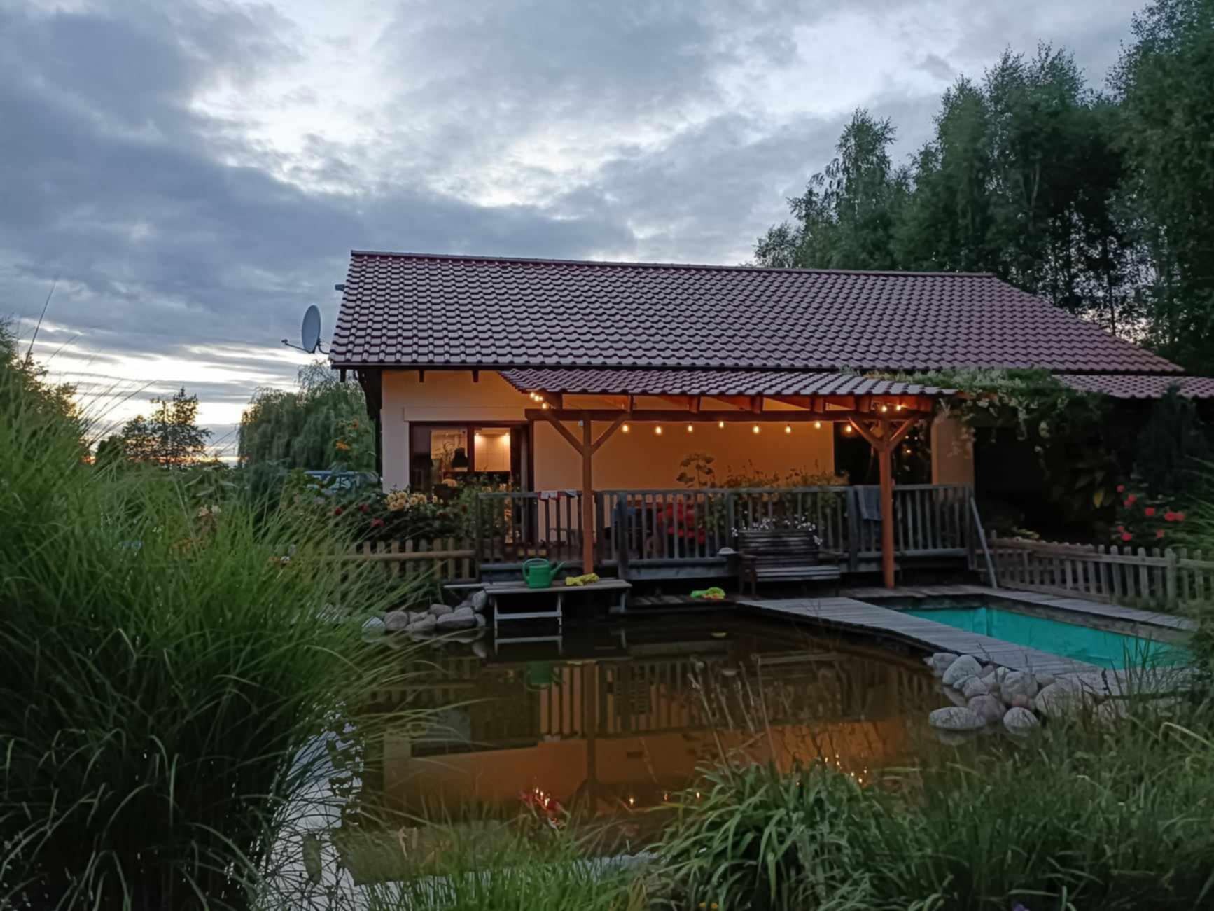 Oświetlona girlandą drewniana konstrukcja przylegająca do domu, z drewnianym tarasem, pomostem nad stawem i małym basenem, widok o zmierzchu.