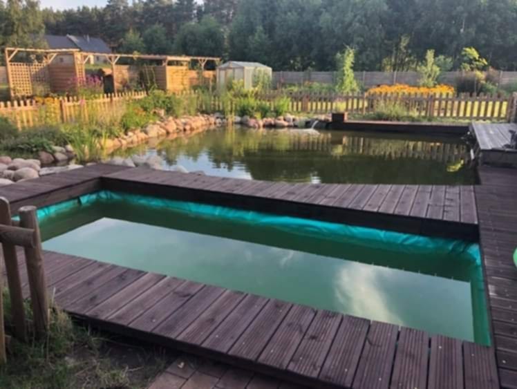 Oczko wodne z drewnianym tarasem i zielonym brzegiem w tle, w ogrodzie z altaną i drewnianym płotem.