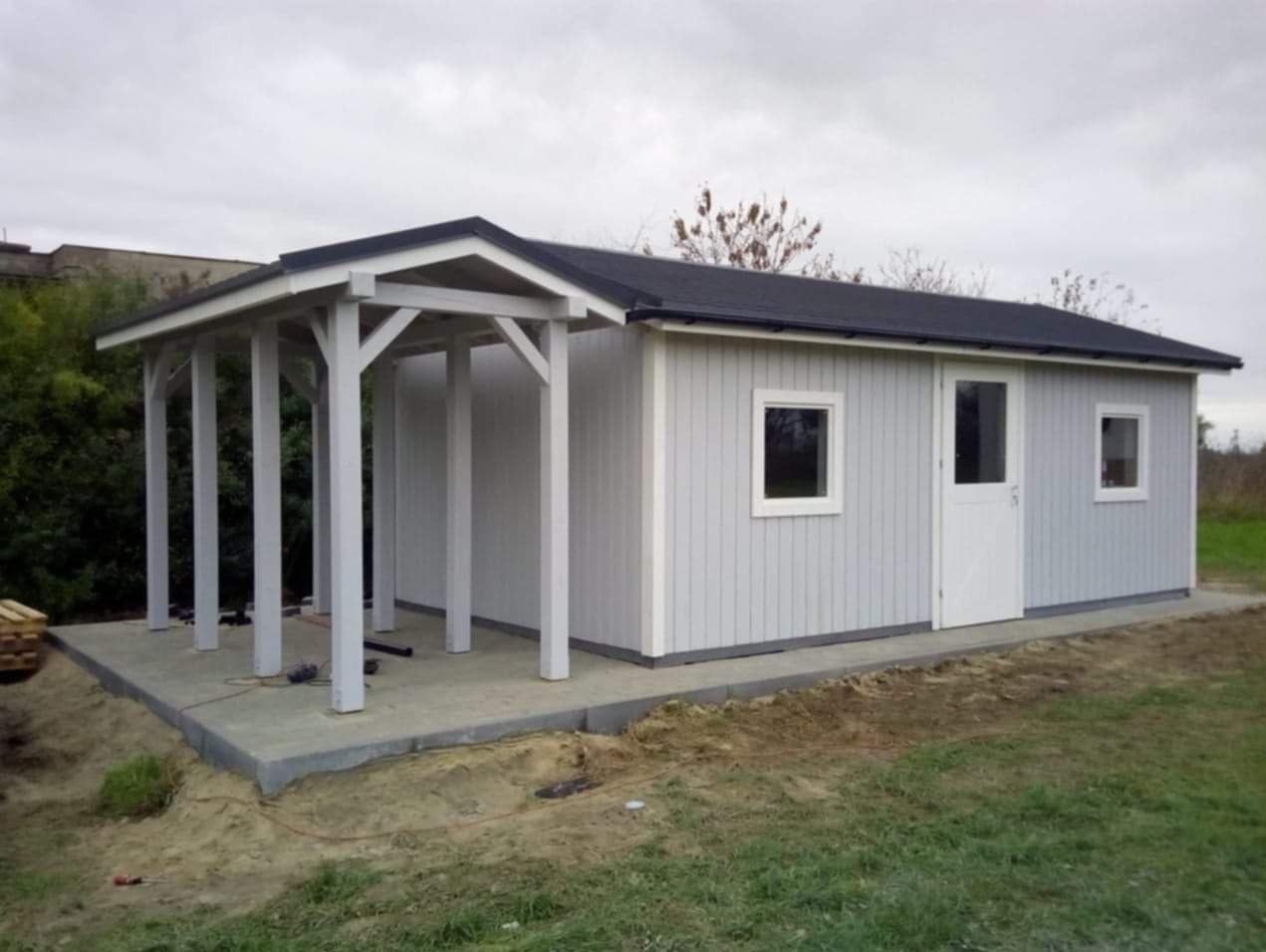 Szara drewniana konstrukcja z zadaszeniem i kolumnami, świeżo postawiona na betonowej wylewce, widoczne narzędzia i kable na podłożu.