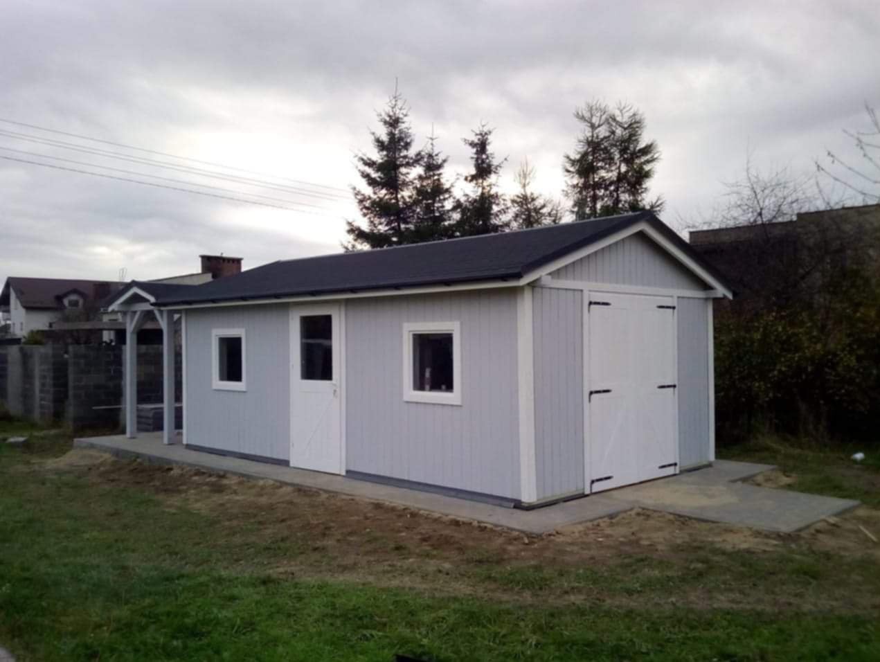 Szara, drewniana konstrukcja z dwuskrzydłowymi drzwiami garażowymi i małą werandą, na betonowej wylewce, w tle drzewa iglaste i domy.