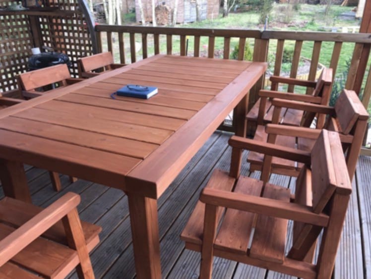 Zestaw mebli ogrodowych z drewna w odcieniach brązu: stół z blatem z desek i sześć krzeseł na drewnianym tarasie z balustradą, na stole leży niebieski notatnik.