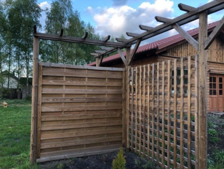 Brązowe ogrodzenie z drewnianych paneli i kraty, połączone z drewnianą konstrukcją pergoli, na tle zielonego trawnika i drewnianego domu z czerwonym dachem.