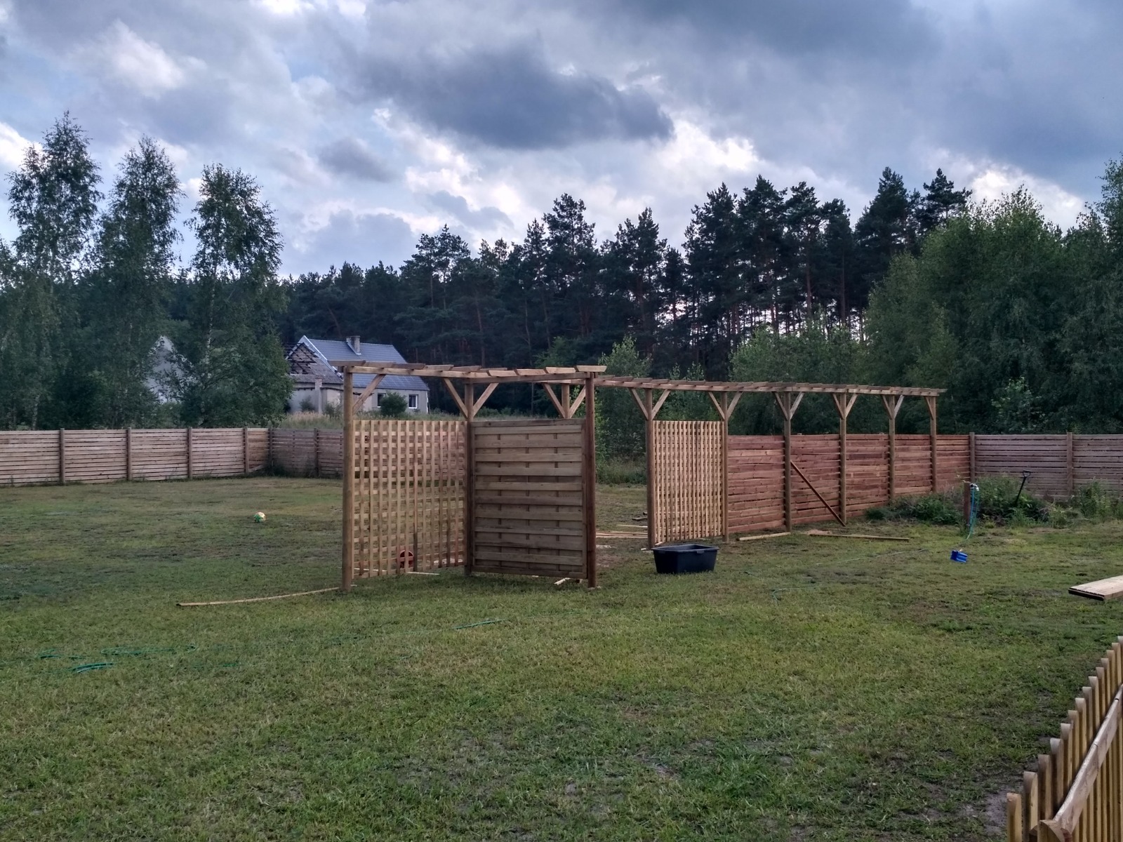 Drewniana pergola z kratownicowymi panelami i ogrodzeniem w tle, widok na posesję z domem pod pochmurnym niebem.