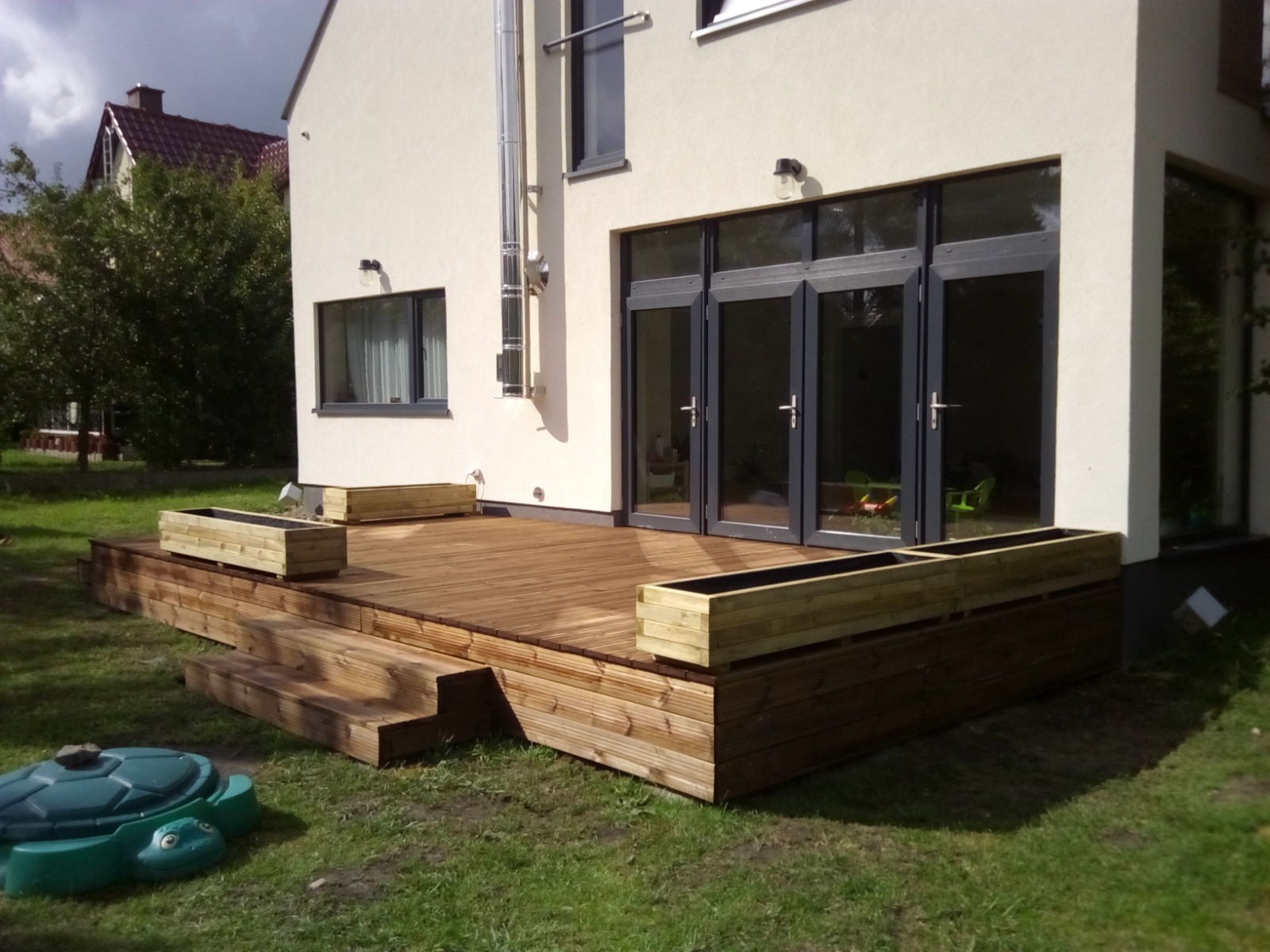 Nowoczesny taras z drewna z drewnianymi donicami, schodkami i widokiem na dom z szarymi ramami okiennymi i drzwiowymi.