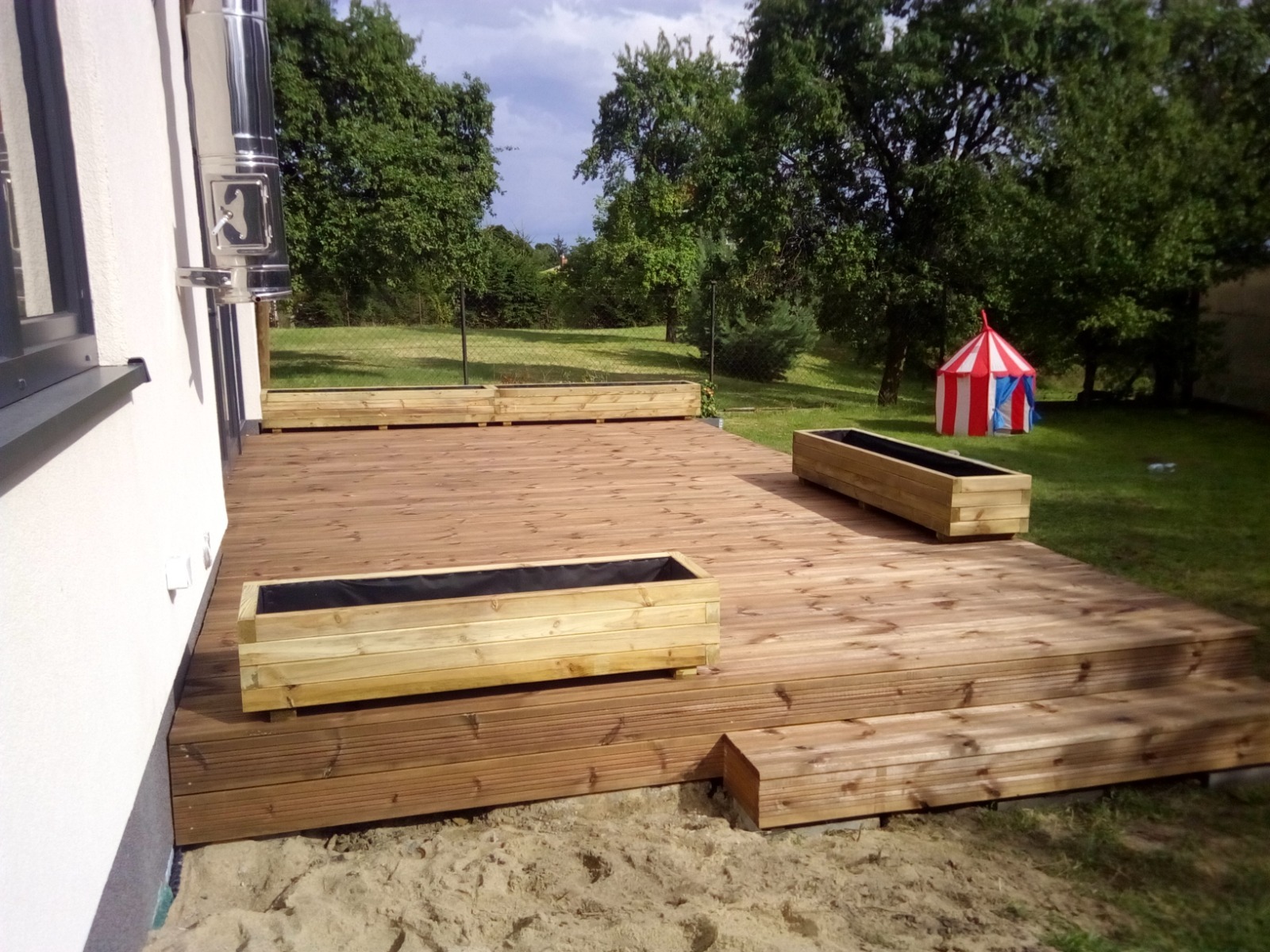 Nowoczesny taras z drewna z jasnymi donicami, na trawniku widoczny namiot cyrkowy.