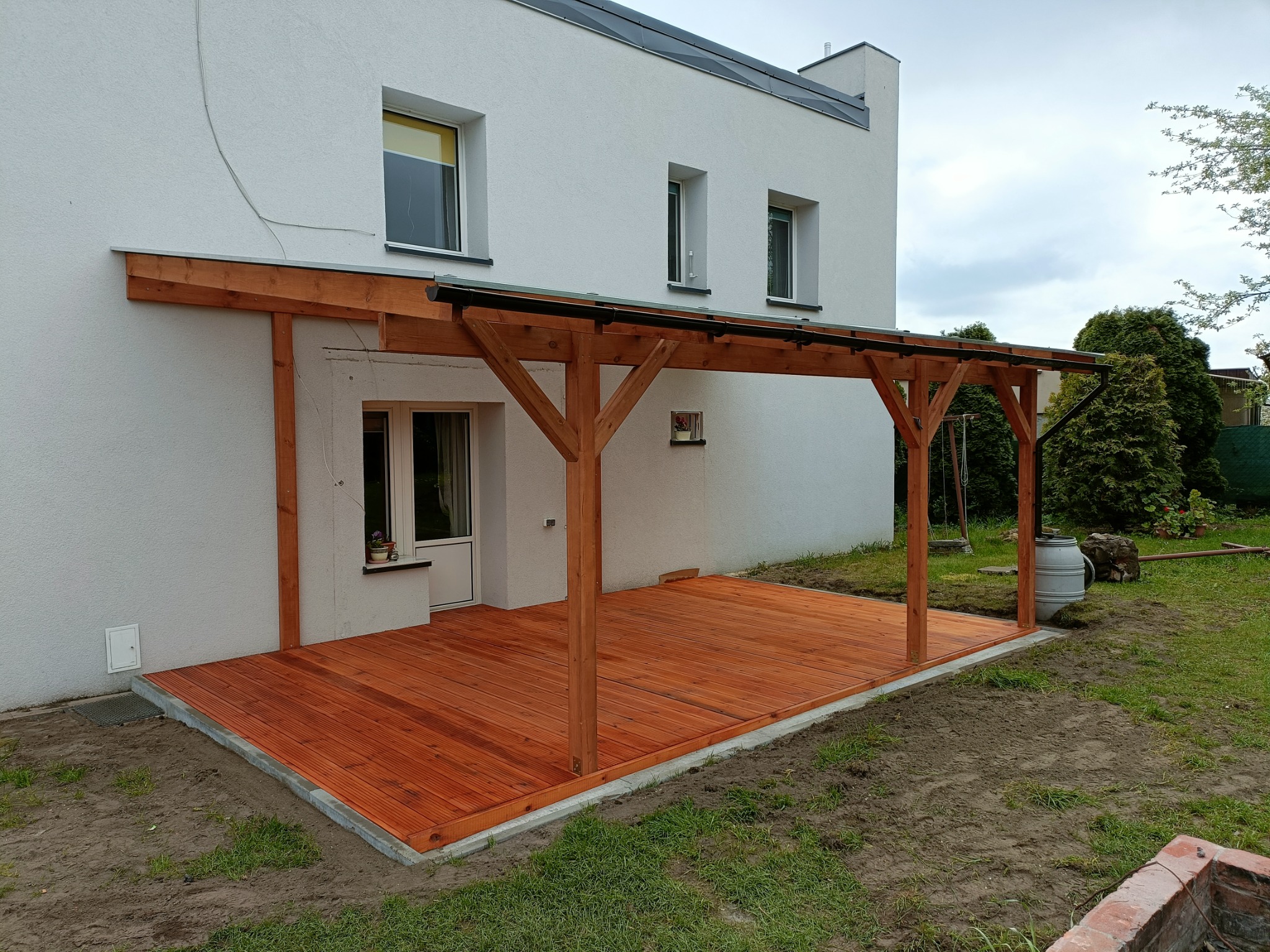 Nowoczesny taras z drewna w kolorze miodowym, zadaszony drewnianą konstrukcją wspartą na słupach, przylegający do białego budynku z widocznymi oknami i zielonym trawnikiem.