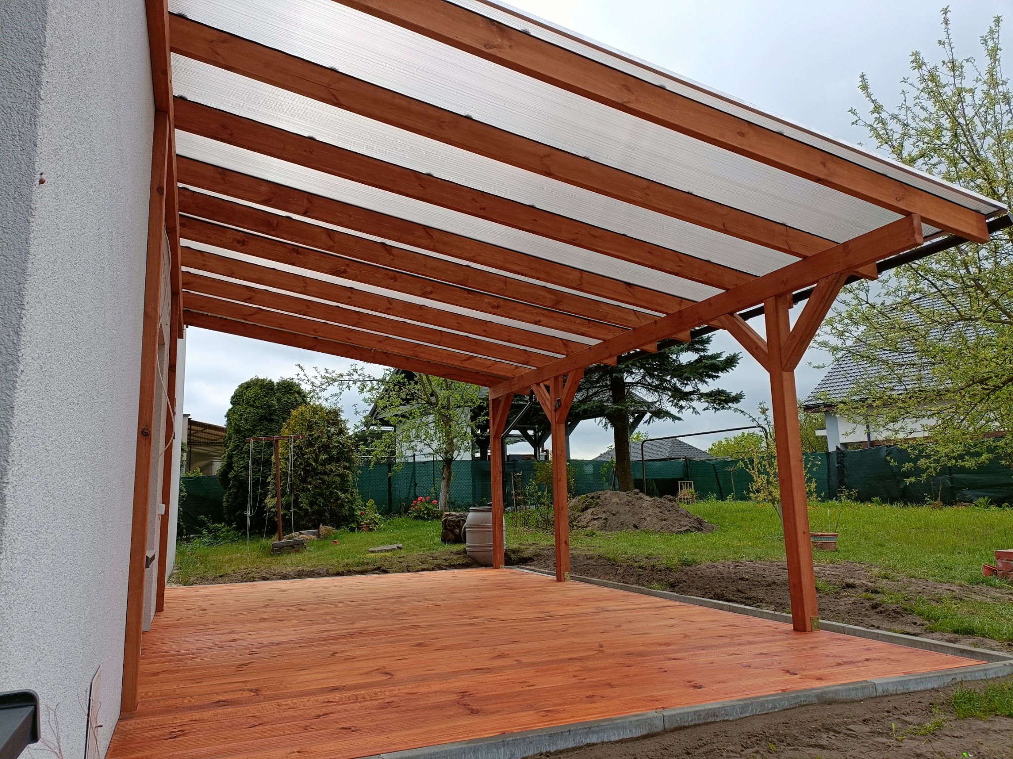 Nowoczesna konstrukcja z drewna i przezroczystego zadaszenia przylegająca do budynku, otoczona zielonym trawnikiem i drzewami.