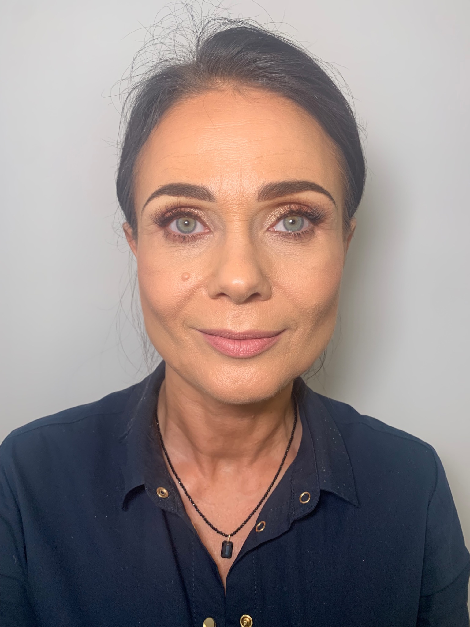 Portret kobiety z delikatnym makijażem, podkreślone oczy z brązowymi cieniami i sztucznymi rzęsami, subtelnie zaznaczone usta, widoczny pieprzyk na policzku.