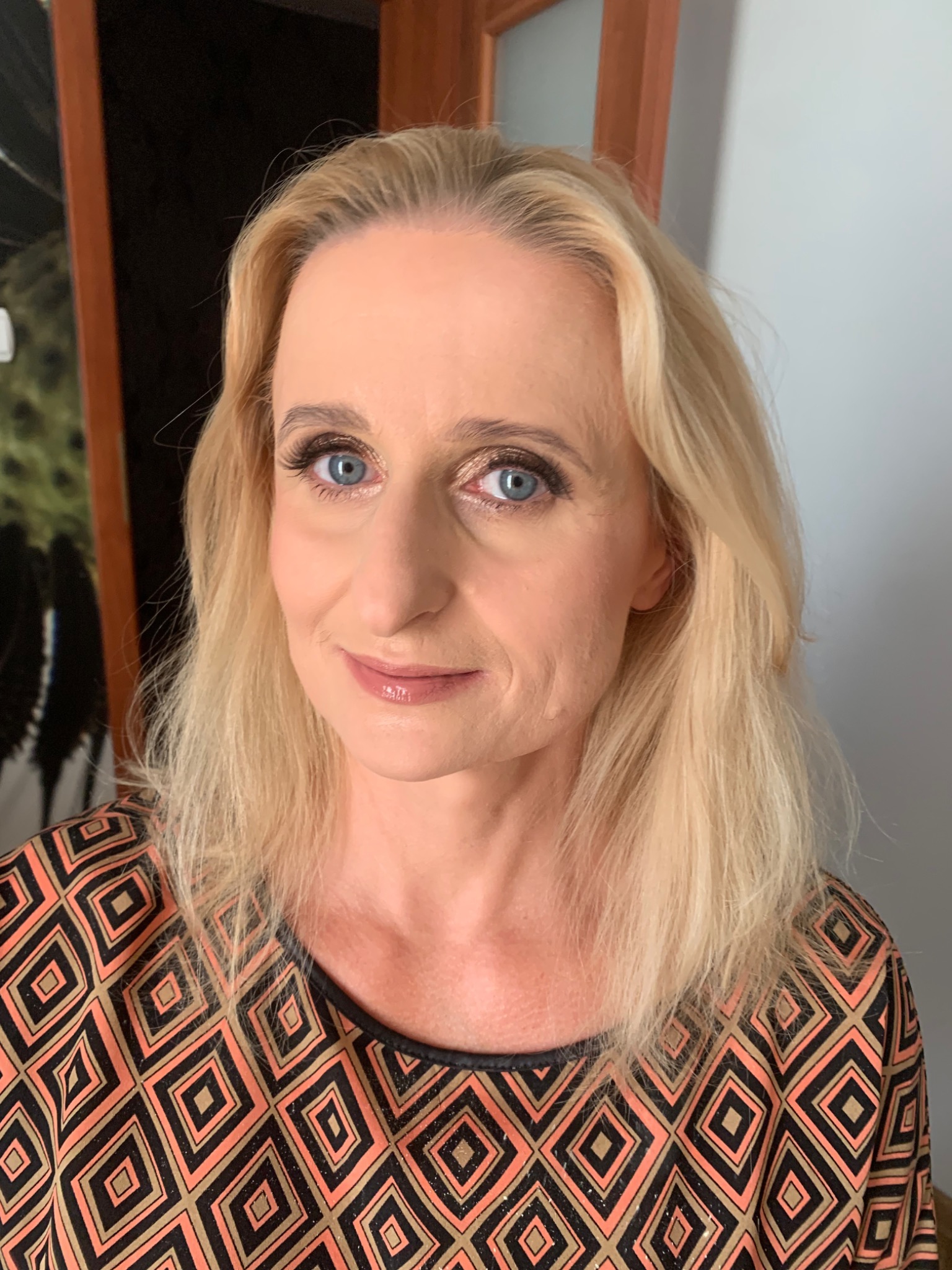 Portret blond kobiety z delikatnym dziennym makijażem, podkreślone oczy i usta, na tle jasnej ściany.