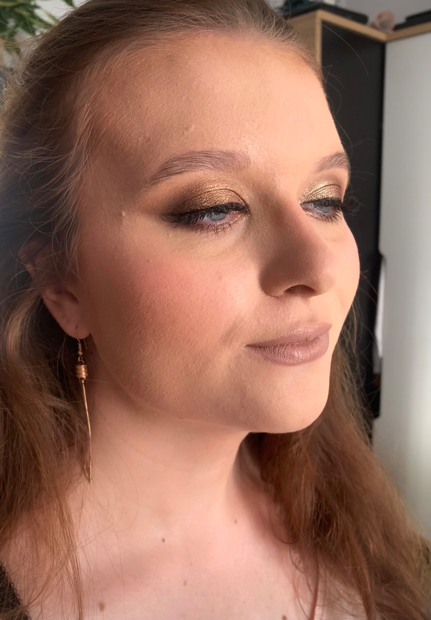 Portret kobiety z profilu z intensywnym, złotym makijażem oczu i delikatnym makijażem ust w odcieniach nude, uzupełnionym długimi, wiszącymi kolczykami.