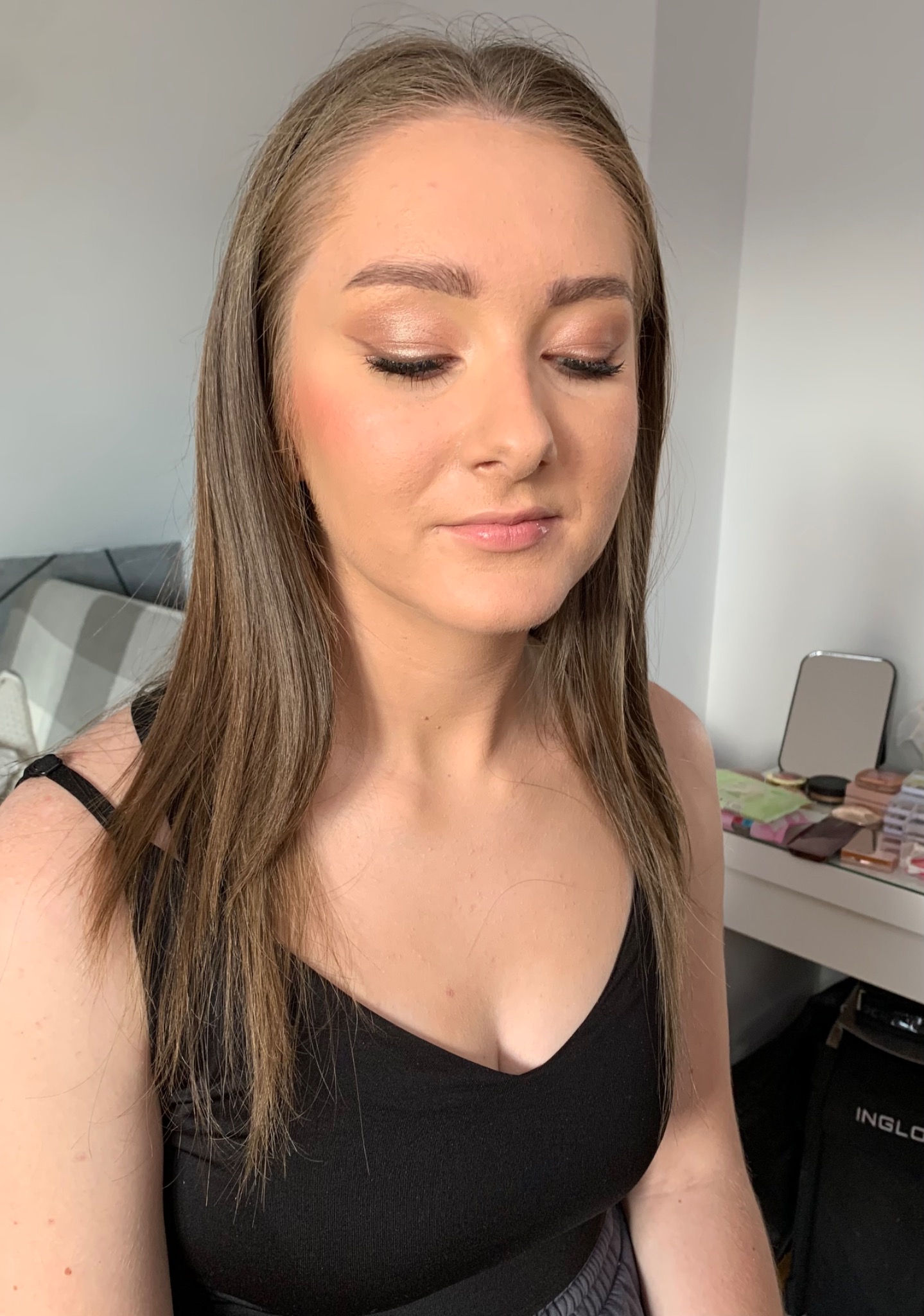 Portret kobiety z delikatnym, błyszczącym makijażem oczu w odcieniach brązu i różu, z zamkniętymi oczami, na tle jasnej ściany i toaletki z kosmetykami.