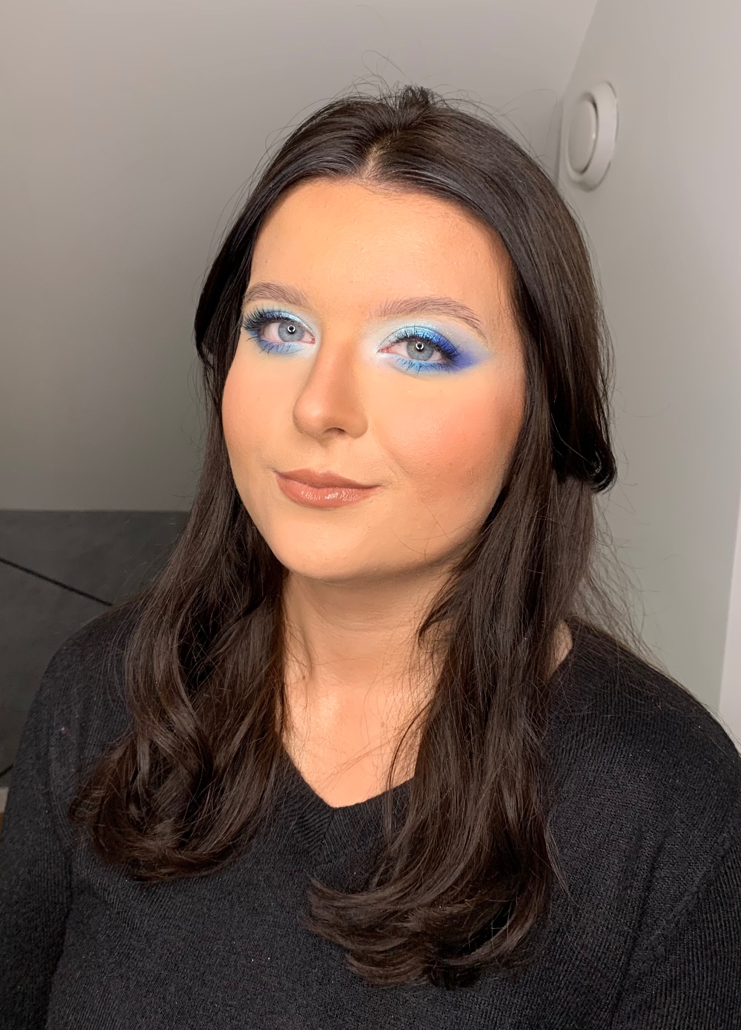 Portret kobiety z intensywnym, niebieskim makijażem oczu z błyszczącymi akcentami i delikatnym, brzoskwiniowym różem na policzkach.