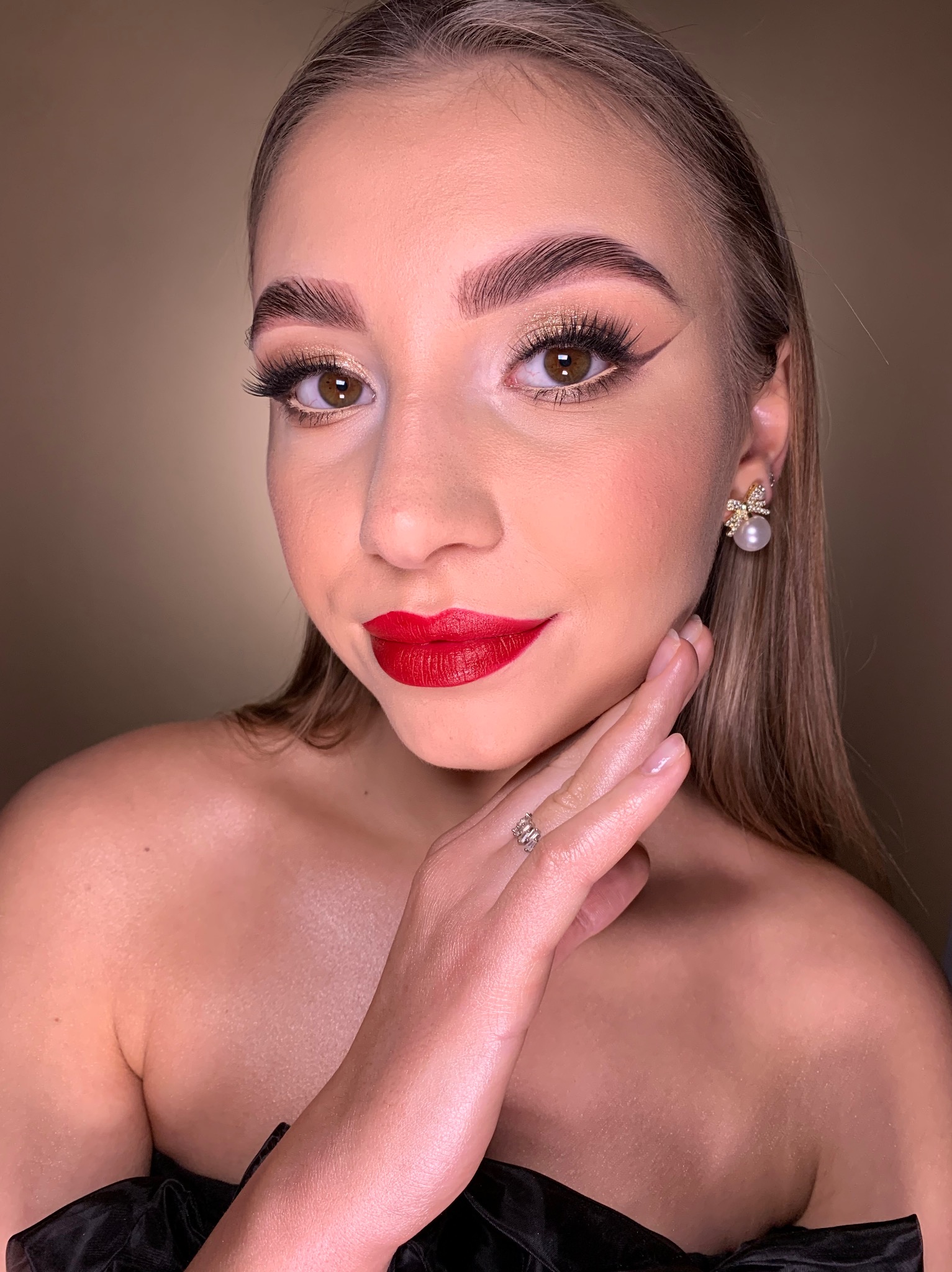Portret kobiety z intensywnym makijażem: czerwone usta, złote cienie z eyelinerem, wyraziste brwi i perłowe kolczyki.