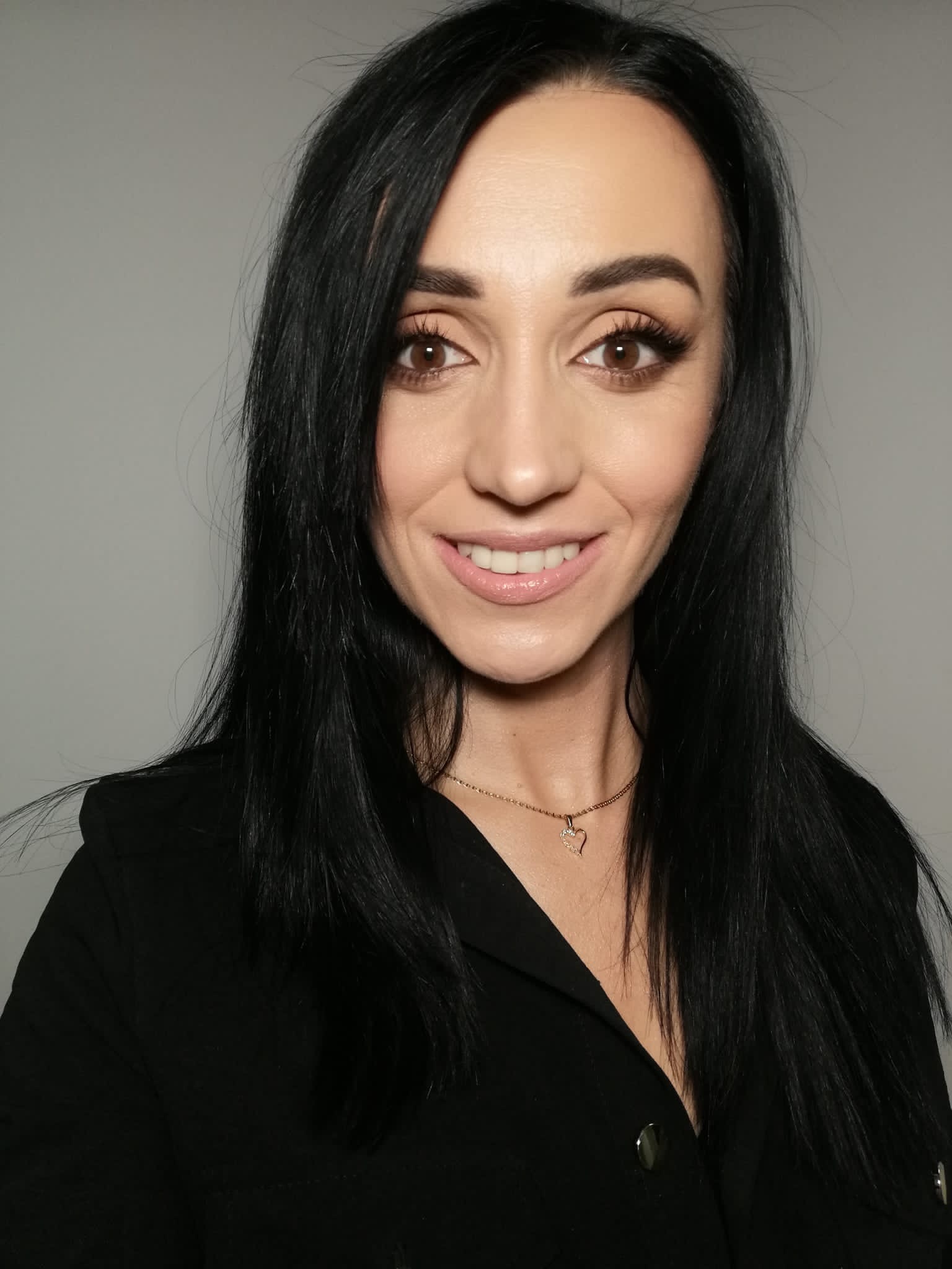 Portret uśmiechniętej brunetki z pełnym makijażem, widoczne sztuczne rzęsy i subtelny błyszczyk na ustach, złoty naszyjnik z zawieszką w kształcie serca.