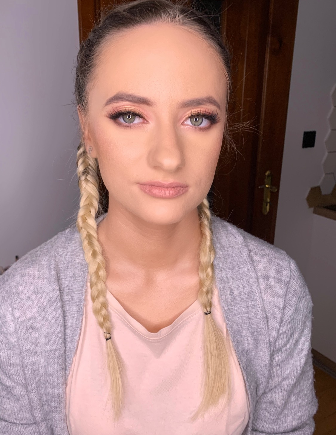 Portret kobiety z delikatnym, brzoskwiniowym makijażem oczu, subtelnym konturowaniem i jasnymi ustami, włosy zaplecione w dwa warkocze.