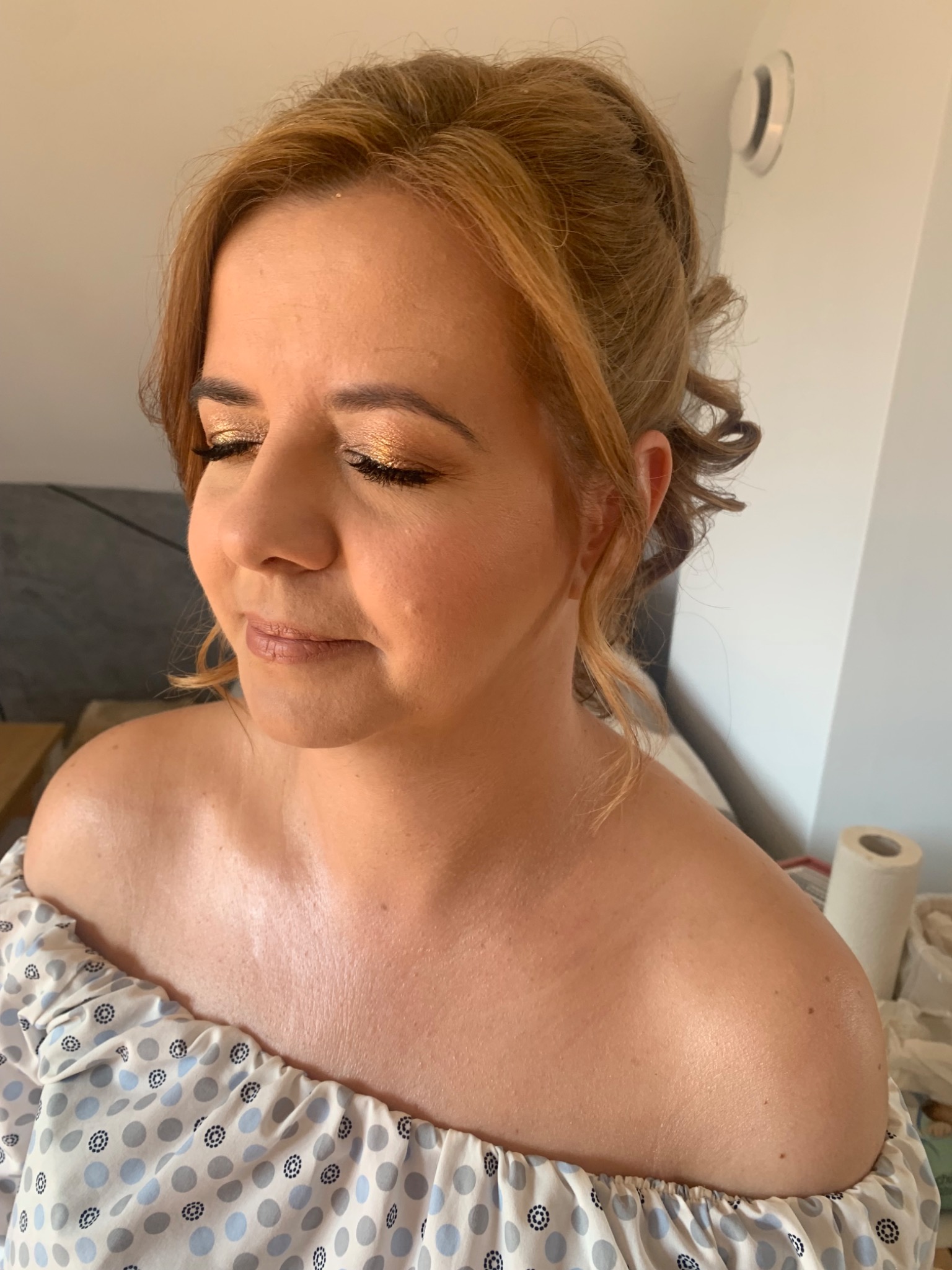 Portret kobiety z zamkniętymi oczami, prezentującej złoty, błyszczący makijaż oczu i delikatny makijaż ust, z upiętymi blond włosami z lokami, w bluzce z odkrytymi ramionami.